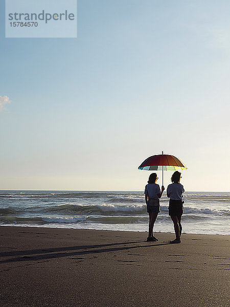 Zwei Frauen mit Sonnenschirm stehen in der Abenddämmerung am Strand  Kedungu Beach  Bali  Indonesien