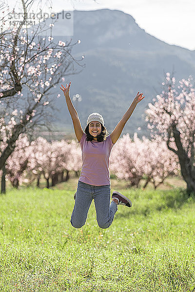 Porträt eines glücklichen Teenagers  der auf einer Wiese vor blühenden Mandelbäumen in die Luft springt