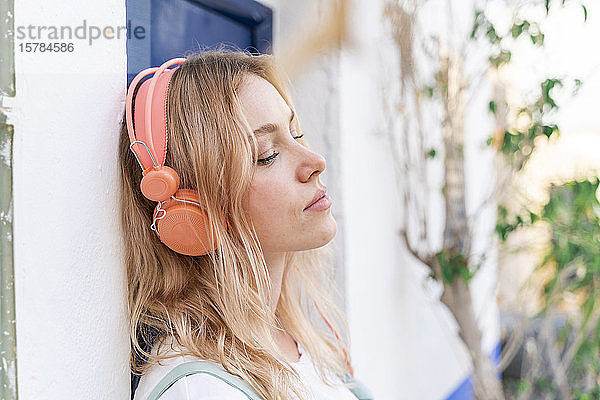 Porträt einer jungen blonden Frau mit geschlossenen Augen  die sich an die Fassade lehnt und mit Kopfhörern Musik hört
