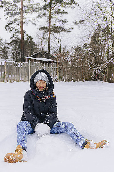 Glückliche Frau sitzt auf Schneefeld und spielt mit Schneebällen