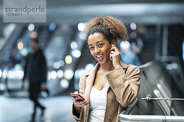 Porträt einer glücklichen jungen Frau mit Handy und Ohrstöpseln in der U-Bahn-Station
