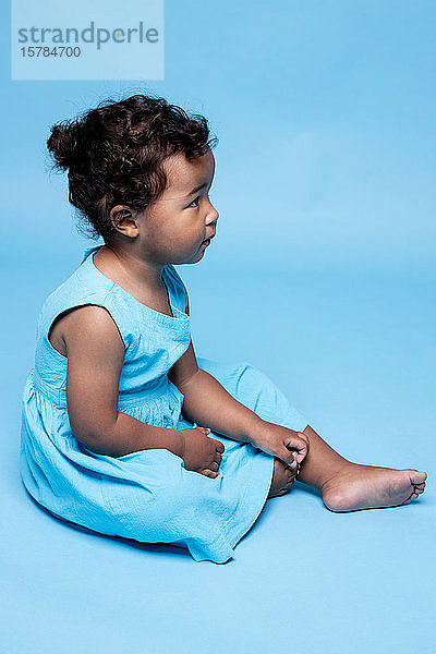 Barfüßiges kleines Mädchen in hellblauem Kleid vor blauem Hintergrund sitzend