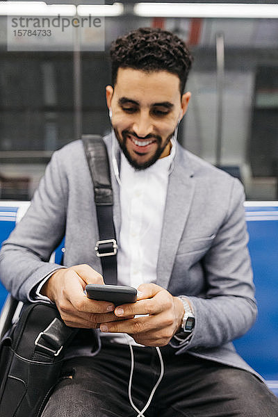 Lächelnder junger Geschäftsmann mit Handy und Kopfhörern in der U-Bahn