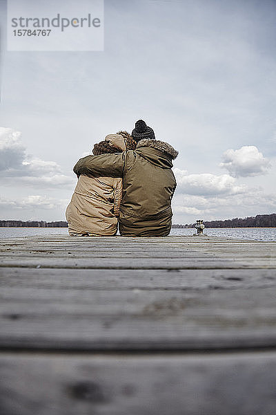 Rückenansicht eines jungen verliebten Paares  das auf einem Steg sitzt und im Winter auf den See schaut