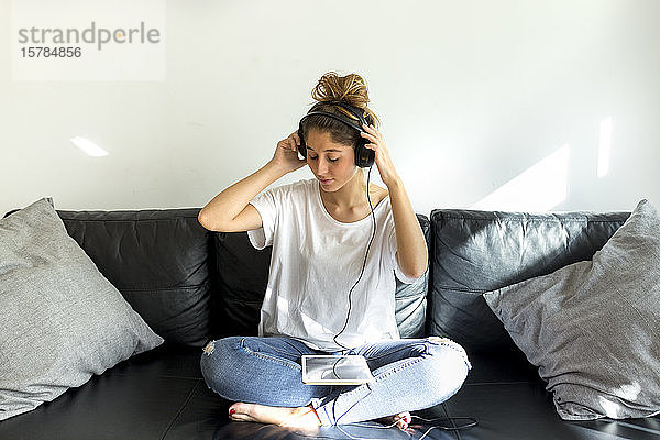 Junge Frau sitzt auf Couch und hört Musik mit Kopfhörern und digitalem Tablet
