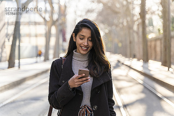 Lächelnde junge Frau mit Smartphone auf Straßenbahnlinie