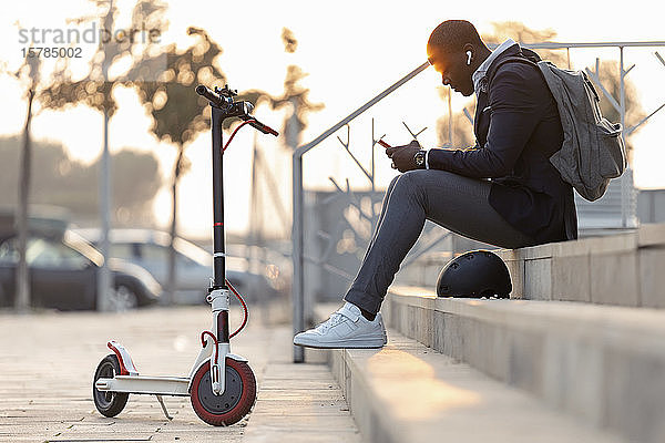 Junger Geschäftsmann mit Schubroller  der bei Sonnenuntergang mit Mobiltelefon und Kopfhörern auf der Treppe sitzt
