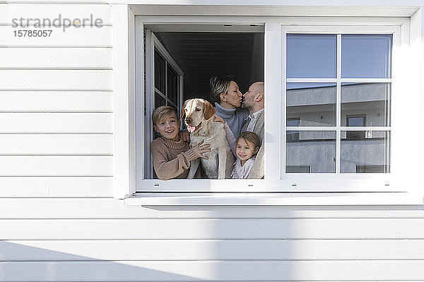 Glückliche Familie mit zwei Kindern und einem Hund am geöffneten Fenster ihres Hauses
