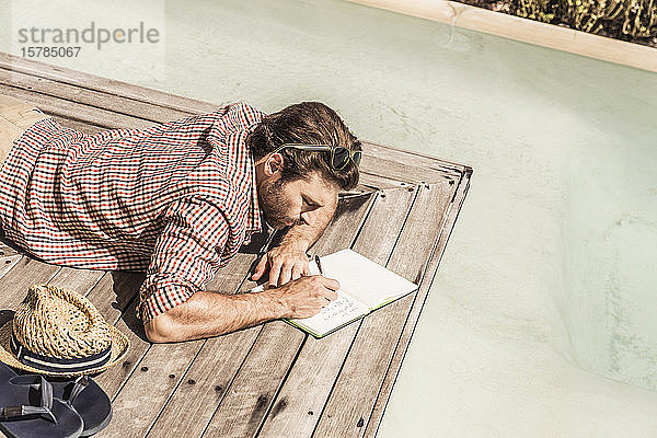 Mann liegt neben einem Pool und schreibt in ein Notizbuch