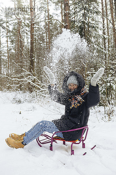 Glückliche Frau sitzt auf Schlitten und wirft im Winterwald Schnee in die Luft