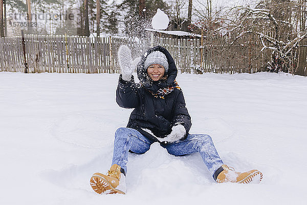 Glückliche Frau sitzt auf Schneefeld und spielt mit Schneebällen