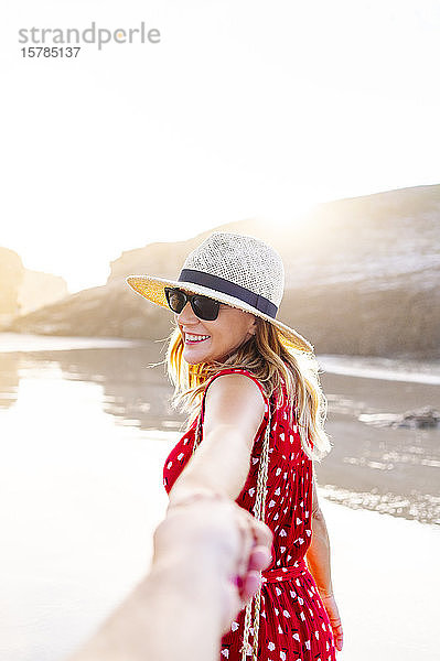 Blonde Frau in rotem Kleid und Hut  die am Strand Hand hält  Naturbogen an der Playa de Las Catedrales  Spanien