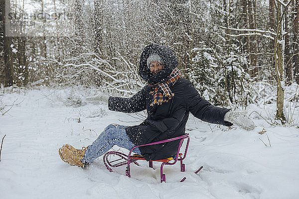 Lächelnde Frau sitzt auf Schlitten und wirft im Winterwald Schnee in die Luft