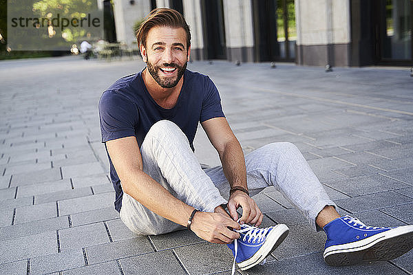 Porträt eines glücklichen Mannes  der in der Stadt auf dem Boden sitzt und seine Schuhe schnürt