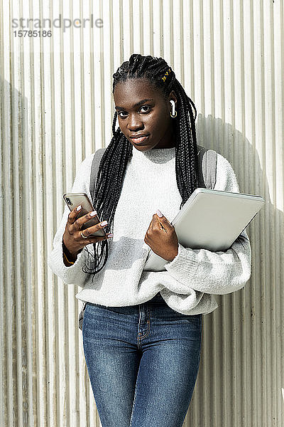 Porträt einer jungen Frau  die mit Handy und Laptop an einer Wand steht