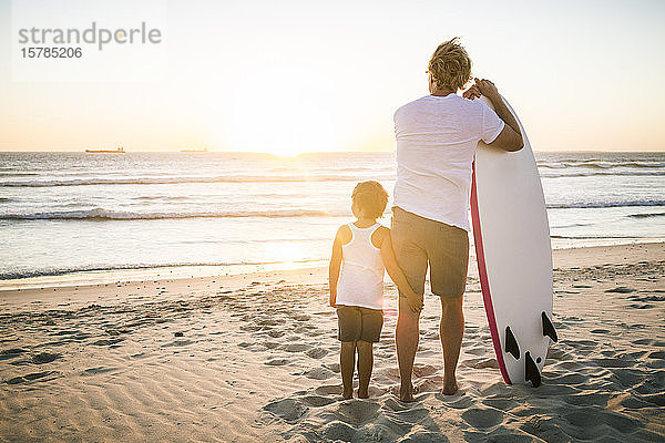 Rückansicht von Vater und Sohn mit Surfbrett  die bei Sonnenuntergang am Strand stehen