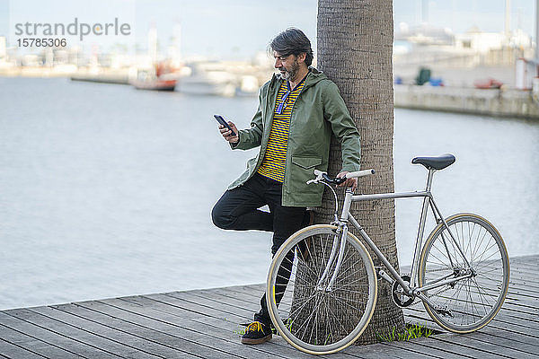 Reifer Mann mit Fixie-Fahrrad  der sich an einen Palmenstamm lehnt und auf sein Handy schaut  Alicante  Spanien