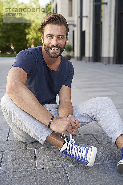 Porträt eines lächelnden Mannes  der in der Stadt auf dem Boden sitzt und seine Schuhe schnürt