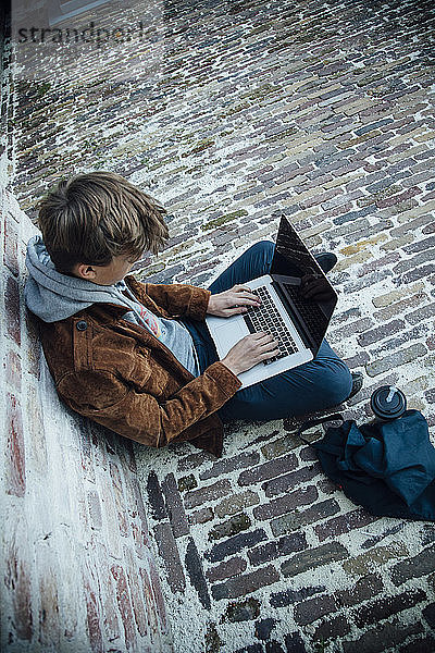 Teenager  der einen Laptop benutzt und auf einem Steinboden in der Stadt sitzt