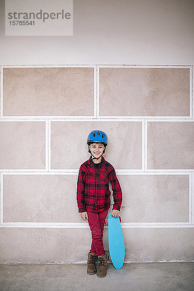Porträt eines lächelnden Jungen mit Helm  der ein Skateboard an einer Wand hält