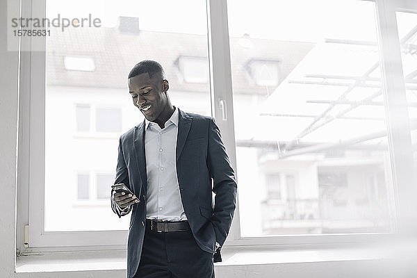 Porträt eines lächelnden jungen Geschäftsmannes  der vor dem Fenster steht und auf sein Handy schaut