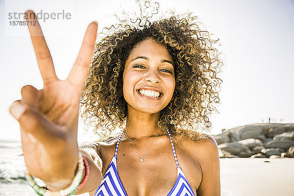 Porträt einer glücklichen jungen Frau beim Siegeszeichen am Strand