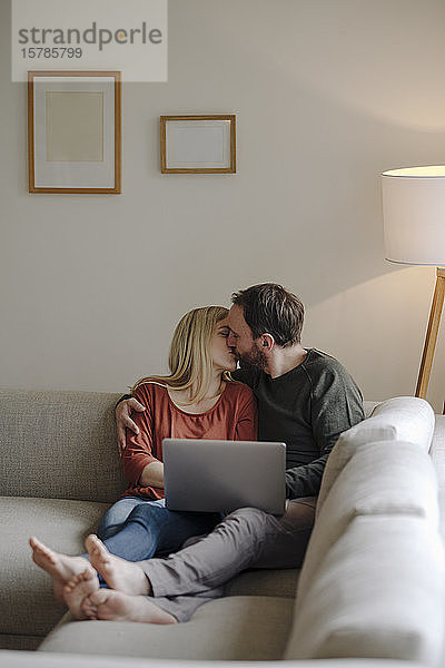 Paar sitzt zu Hause auf der Couch  küsst sich  benutzt einen Laptop
