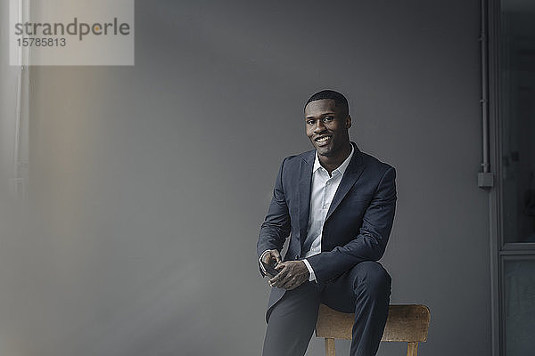 Porträt eines lächelnden jungen Geschäftsmannes mit Smartphone auf Rückenlehne sitzend vor grauem Hintergrund