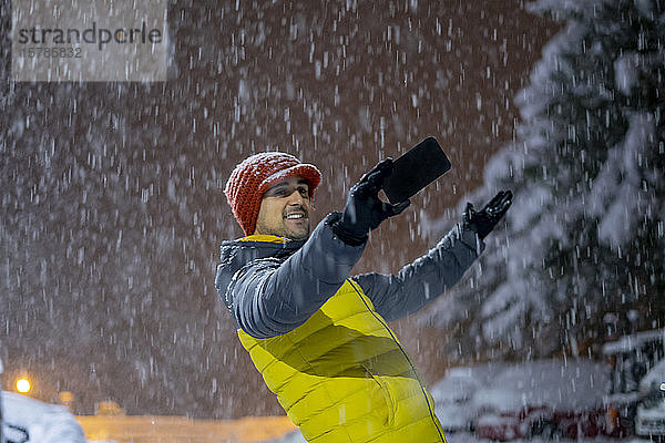 Lächelnder Mann  der sich bei nächtlichem Schneefall ein Selfie nimmt