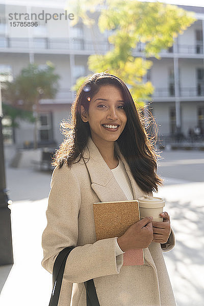 Porträt einer lächelnden jungen Frau mit Buch und Kaffee zum Mitnehmen in der Stadt
