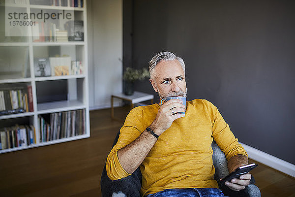 Entspannter reifer Mann zu Hause mit Handy und Tasse Kaffee