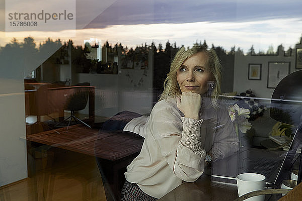 Porträt einer blonden Frau mit Laptop hinter der Fensterscheibe zu Hause