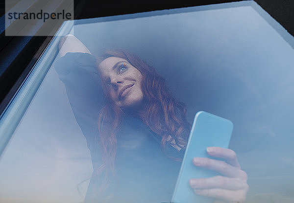 Porträt einer lächelnden rothaarigen Frau  die ihr Handy hinter einer Fensterscheibe hält