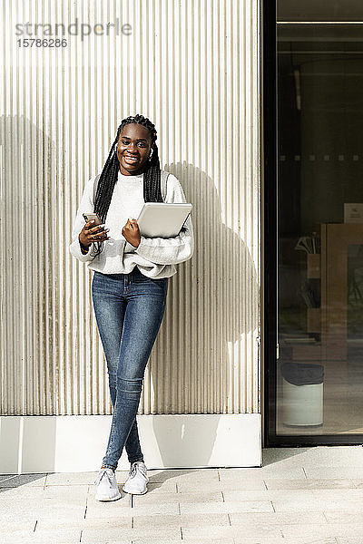 Porträt einer glücklichen jungen Frau  die mit Handy und Laptop an einer Wand steht