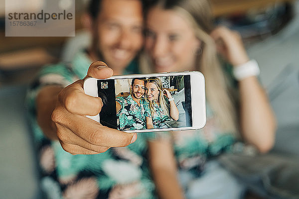 Glückliches Paar sitzt auf der Couch im Wohnzimmer und trägt hawaiianische Hemden und macht einen Selfie