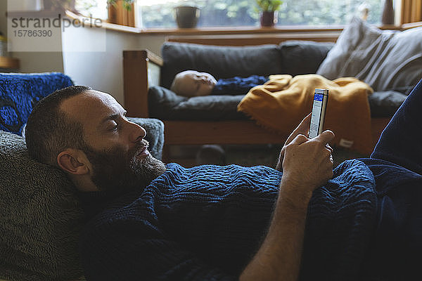Mann überprüft sein Telefon  während sein kleiner Sohn auf dem Sofa schläft