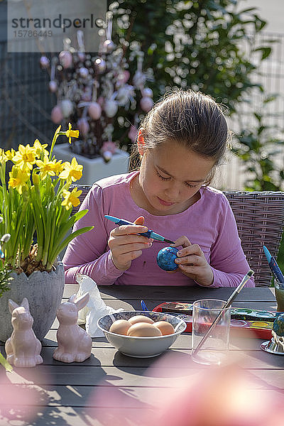 Konzentriertes Mädchen sitzt am Gartentisch und malt Ostereier