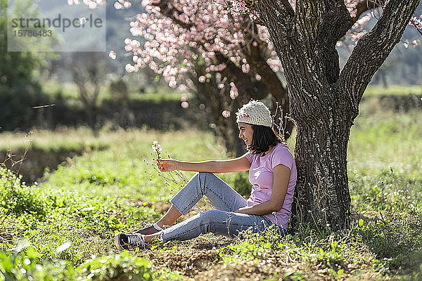 Glückliches Teenager-Mädchen lehnt am Mandelbaum und schaut auf Mandelblütenzweig