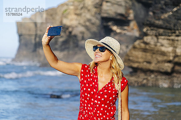 Blonde Frau mit rotem Kleid und Hut  die ein Smartphone benutzt und am Strand ein Selfie macht