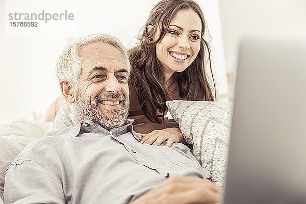 Lächelnder reifer Mann mit Laptop auf der Couch zu Hause  während die Frau zusieht