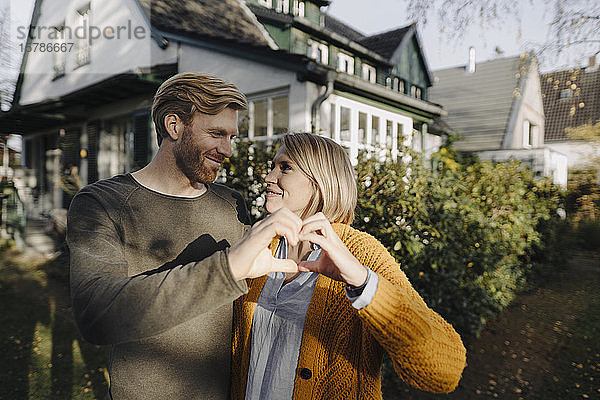 Lächelndes Paar steht vor seinem Haus und formt mit den Händen ein Herz