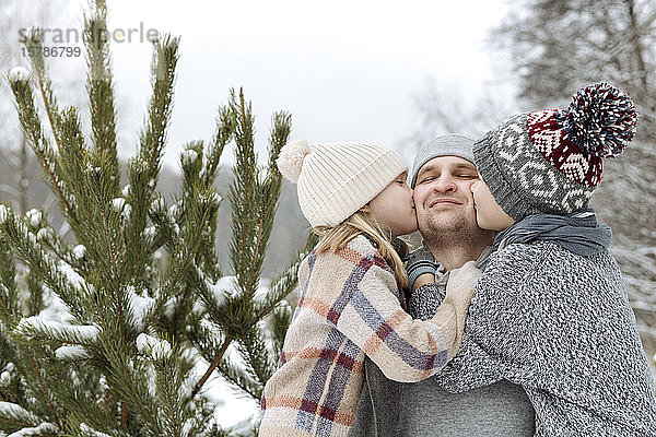 Zwei Kinder küssen im Winter ihren glücklichen Vater an einer Tanne