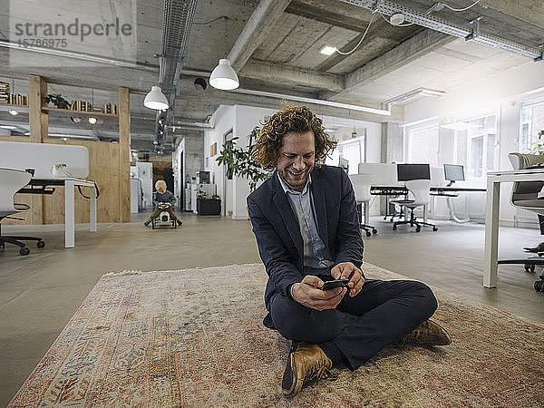 Geschäftsmann sitzt im Büro auf einem Teppich und benutzt sein Handy mit seinem Sohn im Hintergrund
