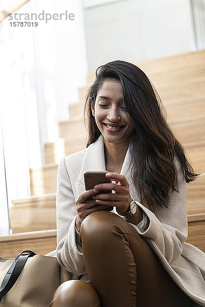 Lächelnde junge Frau sitzt auf der Treppe und benutzt ein Smartphone
