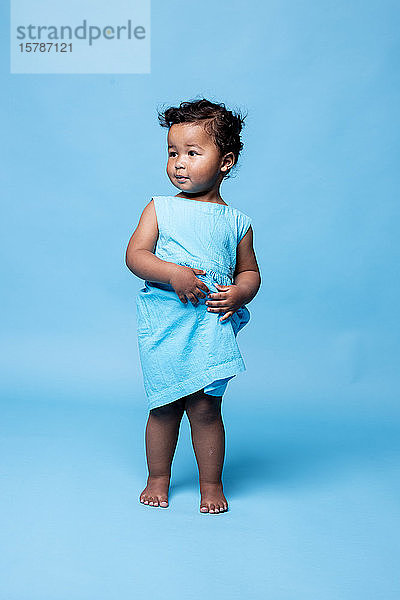 Bildnis eines barfüssigen kleinen Mädchens in hellblauem Kleid vor blauem Hintergrund stehend
