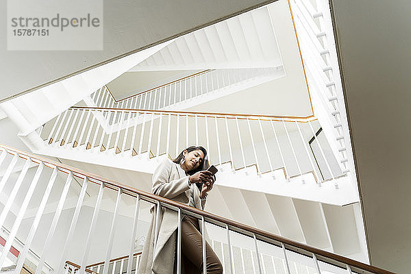 Junge Frau mit Smartphone geht im Treppenhaus die Treppe hinunter
