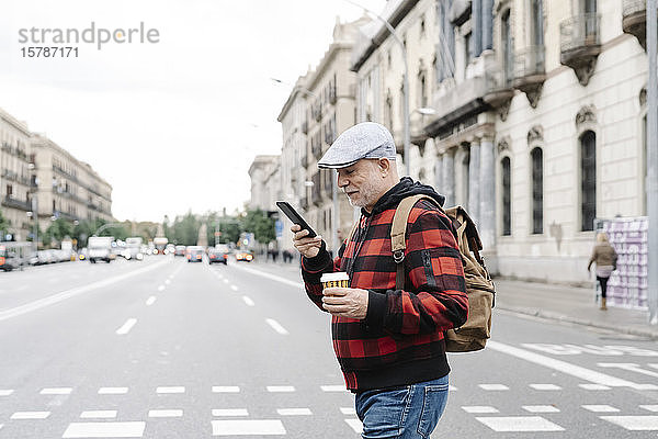 Älterer Mann mit Rucksack und Kaffee  um beim Überqueren der Straße auf sein Handy zu schauen  Barcelona  Spanien