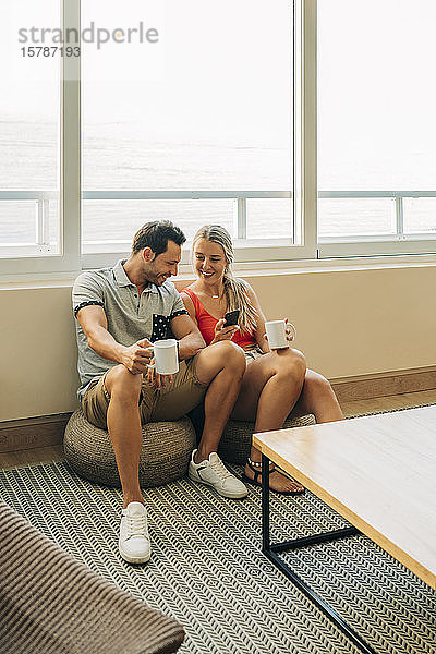 Glückliches Paar sitzt zu Hause am Fenster mit Kaffeetassen und Handy