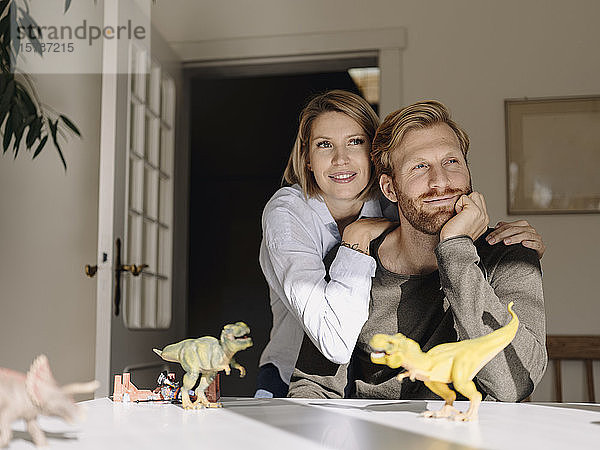 Lächelndes Paar sitzt zu Hause mit Spielzeugdinosauriern am Tisch