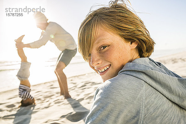 Porträt eines lächelnden Jungen mit dem Vater  der dem Sohn beim Kopfstand am Strand hilft  im Hintergrund